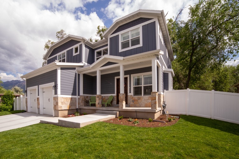 Real daytime photograph of a blue-gray house, showing the full front, roofline, porch and garage with exterior lights off