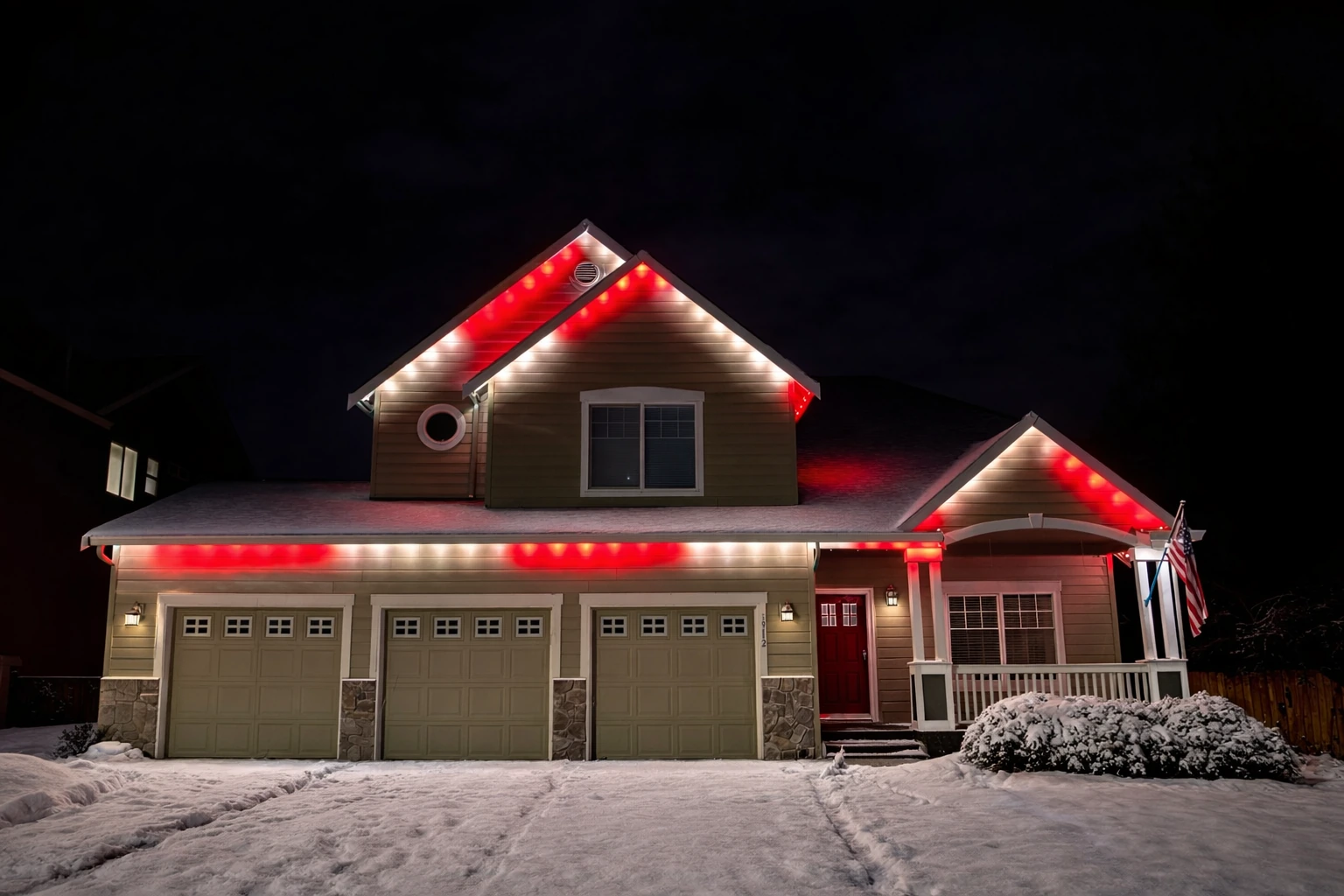 Red and warm-white lighting on a two-story home with snow across the driveway and garden